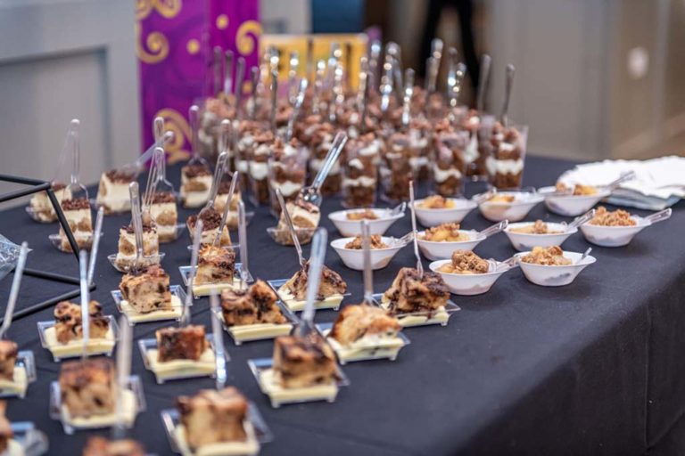 An elegantly arranged dessert table features various individual portions of pastries and desserts in small glass cups and bowls, each with a spoon. The treats are neatly displayed on a black tablecloth, ready to be served.