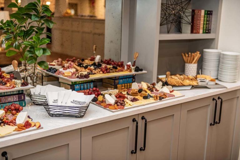 A modern buffet setup featuring a variety of cheeses, fruits, and crackers arranged on wooden charcuterie boards atop books. There are baskets of napkins, stacks of white plates, and breadsticks on the counter, with decorative plants and bookshelves in the background.