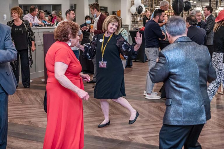 A group of people happily dancing at a social event in a spacious room with wooden floors. A woman in red and a woman in black are notably engaged in the center, while other attendees chat and dance in the background. Black and white balloons are visible.