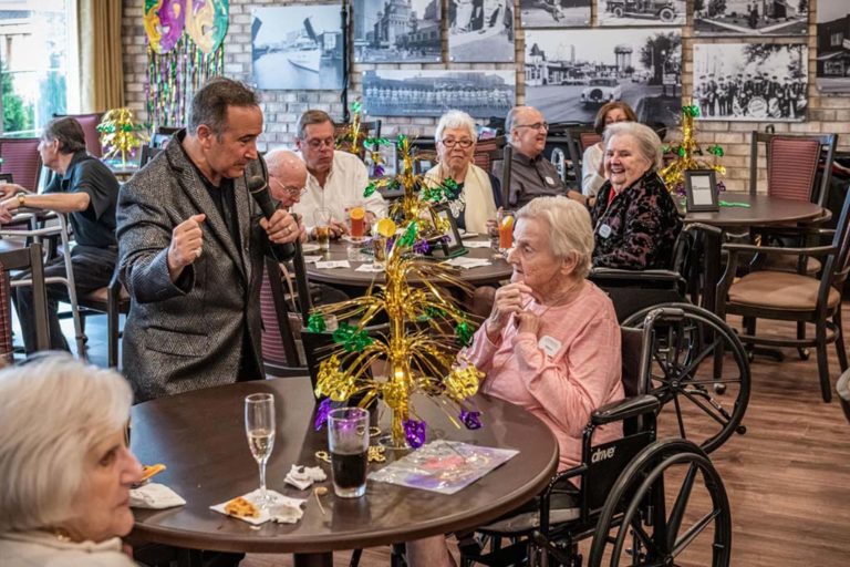 A lively scene of an older people's home dining area with festive decorations. An entertainer engages residents; one woman in a wheelchair converses with him. Other residents sit nearby, smiling and enjoying drinks. The room has a warm, social atmosphere.