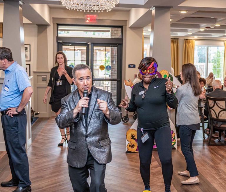 A man in a shiny blazer is singing into a microphone at an indoor event while a woman next to him wearing a colorful mask and black athletic wear dances. Other people in the background are engaged in conversation and activities. The room is well-lit with chandeliers.