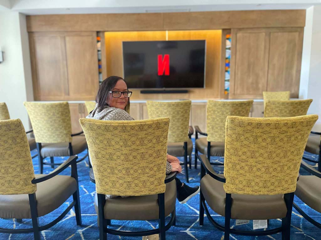 A person sits alone in a small auditorium room on a blue carpet with a geometric pattern. The person is looking back at the camera. Yellow cushioned chairs, a large TV screen with the Netflix logo, and wooden paneling are visible.