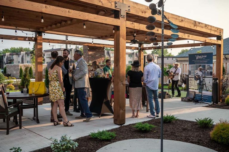 A group of people is gathered under a wooden pergola at an outdoor event. They are socializing and listening to live music performed by a band in the background. Some are standing by cocktail tables, and the atmosphere is relaxed and casual.