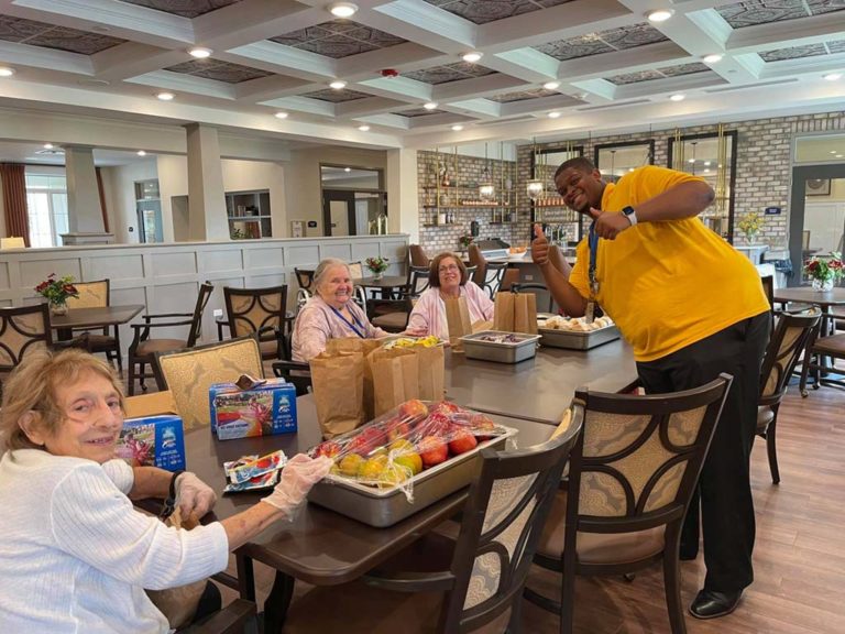 A man in a yellow shirt stands behind a table in a spacious, well-lit room. Three elderly women are seated at the table, engaged in packing bags with food items. The mood appears cheerful, with the man giving a thumbs-up gesture.