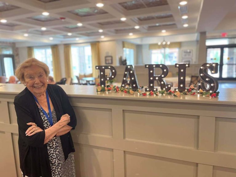 An elderly woman stands smiling with arms crossed in front of a sign that spells "PARIS" in large, illuminated letters. The letters are decorated with small lights and flowers. The setting appears to be inside a well-lit room with a coffered ceiling and large windows.