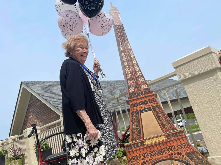 An elderly woman smiling and holding a cluster of balloons stands next to a cardboard cut-out of the Eiffel Tower. She is outside on a sunny day, wearing a black cardigan over a floral dress. The background shows a building, shrubbery, and a clear blue sky.