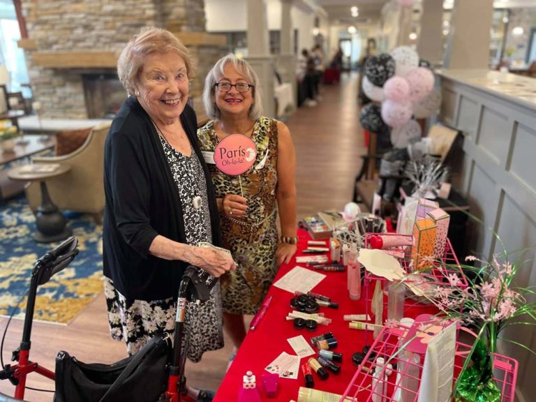 Two elderly women at a festive event are standing next to a table displaying various beauty products. One woman is holding a "Paris" sign. The backdrop features a warmly lit room with comfortable seating, flowers, and balloons, creating a lively atmosphere.