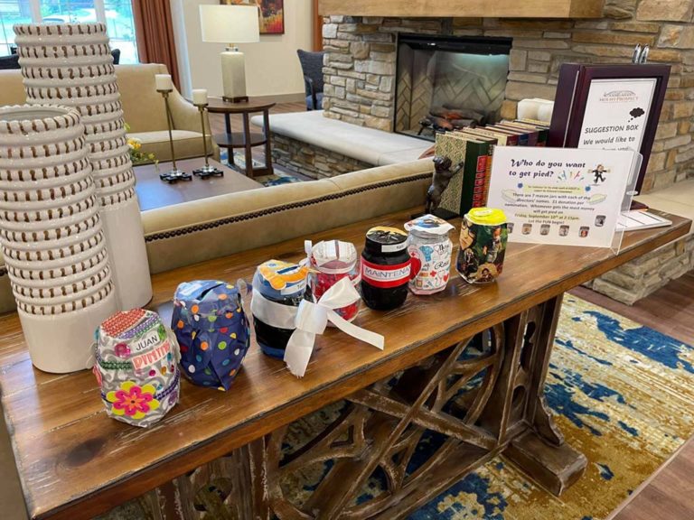 A wooden table displays several uniquely decorated jars, each with colorful designs, ribbons, and labels. A nearby sign titled "Suggestion Box" includes a voting section with text and small images. The setting features a cozy, well-decorated room with a fireplace.