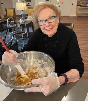 An elderly woman with short, light brown hair and glasses is smiling while mixing ingredients in a large metal bowl. She is wearing a black top and plastic gloves. In the background, there is a walker, a lamp, and colorful furnishings.