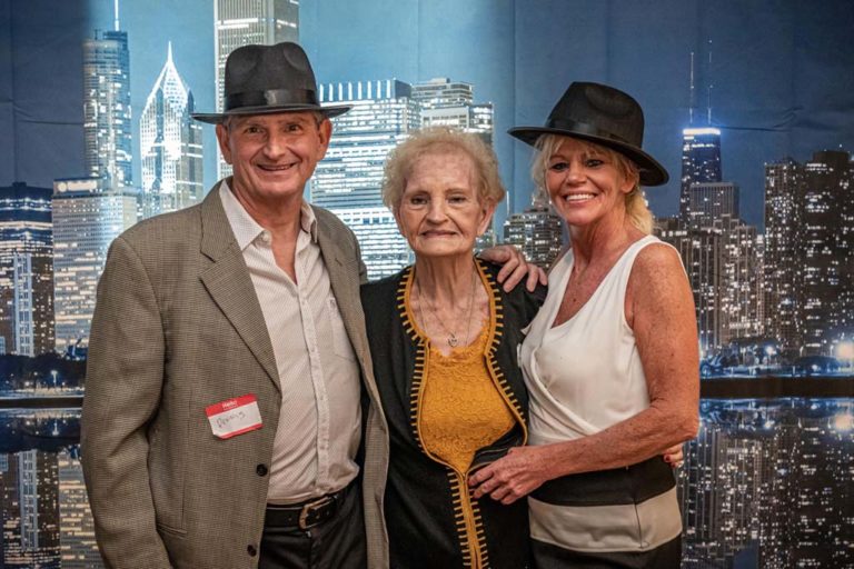 Three people stand together in front of a city skyline backdrop at night. The person on the left is wearing a gray jacket and hat, the person in the middle is older and wearing a yellow and black outfit, and the person on the right is wearing a white top and black hat. All are smiling.