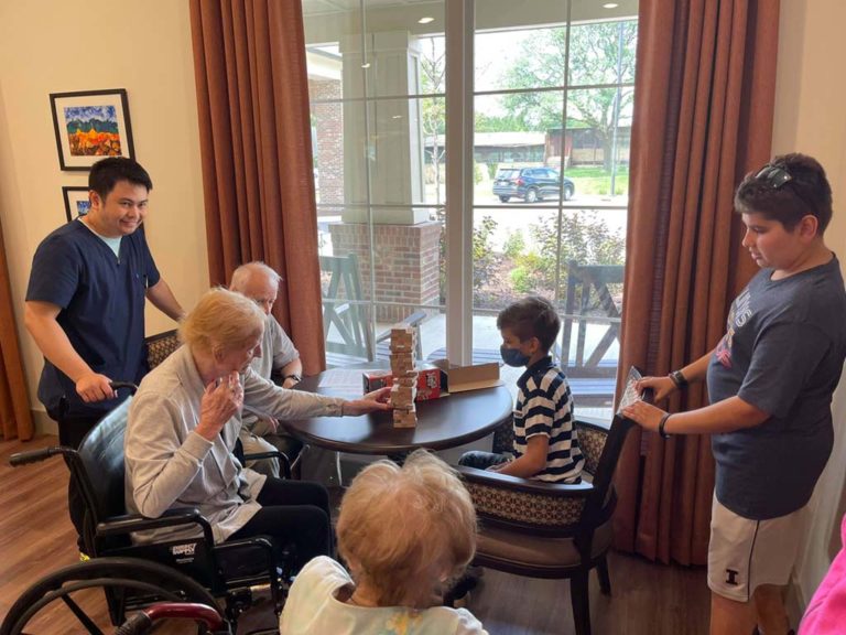 A group of elderly individuals and children are gathered around a table, playing a game of Jenga. One elderly woman in a wheelchair is reaching to place a block. A young boy in a striped shirt is focused on the game. A caregiver and another child are observing.