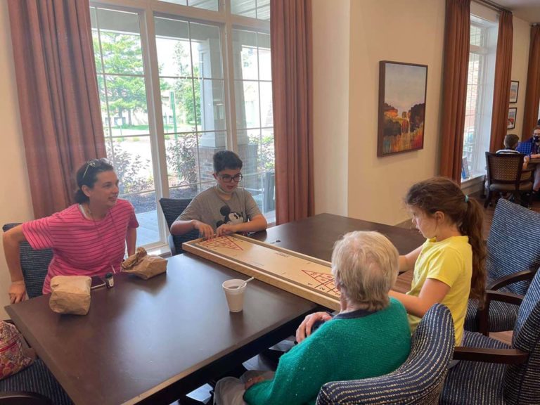 Four people, a senior woman, an adult woman, and two children, are seated around a table in a well-lit room with large windows. They appear to be playing a board game and have snacks and a drink on the table. The room has comfortable chairs and a painting on the wall.