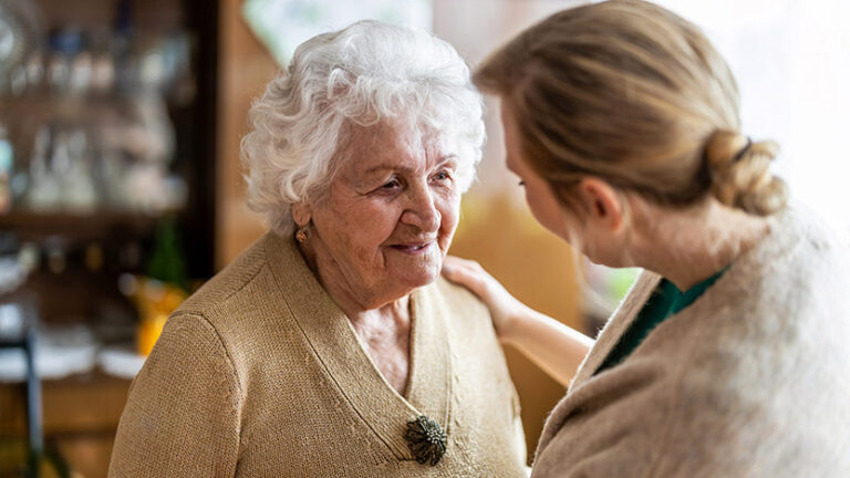 An elderly woman with curly white hair wearing a beige cardigan smiles warmly at a younger woman with light brown hair pulled back, who is gently touching her shoulder in a comforting gesture. The setting is a cozy indoor space.