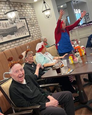 Elderly people gather in a festive setting. Some wear pumpkin headbands and costumes. One person raises their arms enthusiastically. Tables are decorated with Halloween-themed items. They appear to be in a communal area with bright lighting.