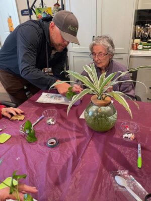 A man helps an elderly woman with a craft project at a table covered with a purple cloth. They are surrounded by art supplies and a potted plant. The setting appears to be a cozy room with a bookshelf in the background.