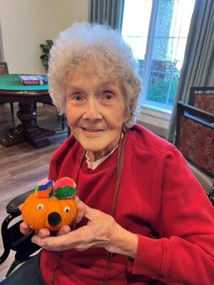 An elderly woman with curly white hair and a red sweater smiles while holding a small pumpkin decorated with colorful pom-poms and googly eyes. She is sitting in a cozy room with a table and window in the background.