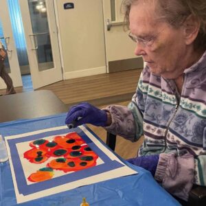 Elderly woman wearing gloves and a patterned sweater creates abstract art on paper using various colors, seated at a table covered with a blue cloth.