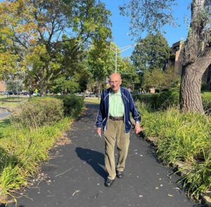 An elderly man is walking on a paved path in a park, surrounded by trees and bushes. The sky is clear and sunny, and shadows are cast on the ground. The man is wearing a dark jacket, green shirt, and brown pants.