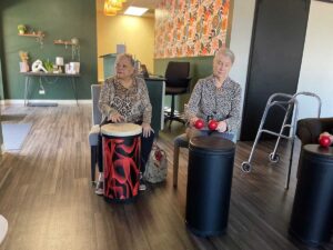 Two women in a brightly lit room sit on chairs playing drums. One woman is holding drumsticks, and the other taps with her hands. A walker stands nearby, and the room features wooden floors and patterned walls.