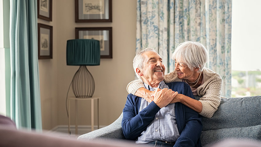 Senior woman hugging senior man while he is sitting on couch in living room