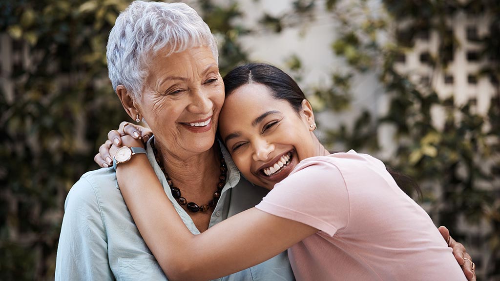 An elderly woman with short white hair and a younger woman with long dark hair share a warm embrace outdoors. Both are smiling joyfully. The elderly woman wears a light blue blouse, and the young woman has on a pink shirt. Green foliage is in the background.