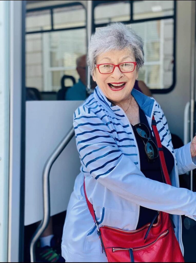 Elderly woman with short grey hair and red glasses, wearing a blue and white striped jacket, smiles while holding onto a metal handrail. She has a red bag across her shoulder and stands in front of a doorway.