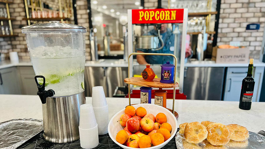 A countertop with a variety of refreshments including a water dispenser with cucumber slices, cups, a bowl of peaches, a plate of pastries, and a popcorn machine with condiments in front of it. A wine bottle is placed on the right side.