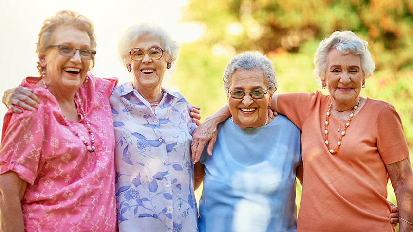 Four elderly women are standing side by side outdoors, smiling joyfully with their arms around each other. They are dressed in colorful clothing, displaying a sense of camaraderie and happiness. The background shows greenery, suggesting they are in a garden or park.