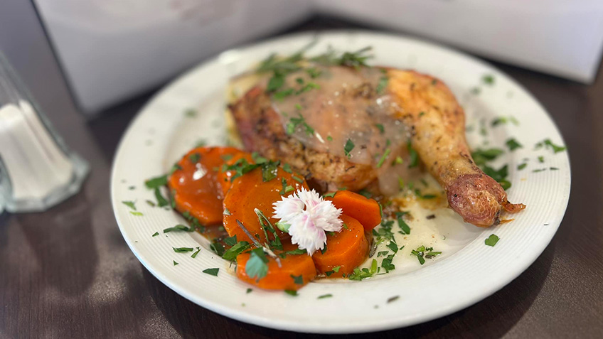 A plated dish features a roasted chicken leg topped with gravy, accompanied by glazed carrot slices. The plate is garnished with fresh herbs and decorated with two small white flowers.