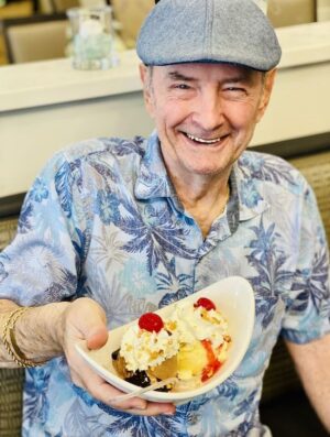 A smiling man wearing a blue tropical shirt and a grey flat cap is holding a bowl with a dessert that includes ice cream, whipped cream, and cherries. He is seated indoors, possibly in a restaurant.
