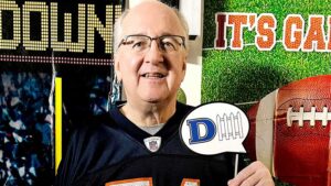 A smiling middle-aged man wearing glasses and a black football jersey. He is holding a speech bubble sign with the letter "D" and a representation of a picket fence, symbolizing defense. Banners in the background include the word "Touchdown" and a football image.