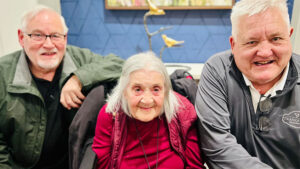 Three older adults are posing together indoors. The person on the left is wearing glasses and a green jacket, the person in the center has long white hair and is wearing a red top, and the person on the right has short white hair and is wearing a dark gray shirt.