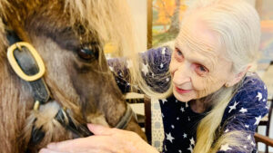 An elderly woman with long white hair and a star-patterned shirt gently pets a small horse's face indoors. She is smiling, and the horse is calm and close to her. A painting is visible in the background.