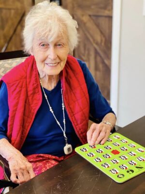 An elderly woman with short white hair, wearing a blue shirt and a red vest, is sitting at a table and smiling. She is holding a green bingo card and appears to be playing bingo. There is a wooden door in the background.