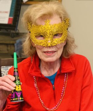 An elderly woman with light hair is dressed in a red jacket and wearing a decorative, gold masquerade mask. She holds a colorful noisemaker and has pink beaded necklaces around her neck, suggesting she is celebrating a festive occasion.