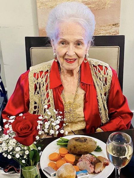 An elderly woman with white hair, dressed in a red and gold outfit, sits at a table. In front of her is a meal with a potato, vegetables, and meat, along with a glass of water. A vase containing a red rose and white baby's breath flowers is also on the table.