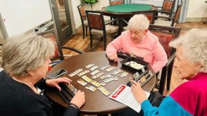 Three elderly women are seated around a circular table, deeply focused on a game involving numbered tiles. They are in a cozy room with a poker table and chairs in the background. One woman is reading game instructions while the others arrange their tiles.