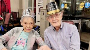 An elderly couple is sitting together and smiling at the camera, both wearing festive hats. The woman has a "Happy New Year" headband on, and the man is wearing a "Happy New Year" top hat. They are holding hands, celebrating in a cozy indoor setting.