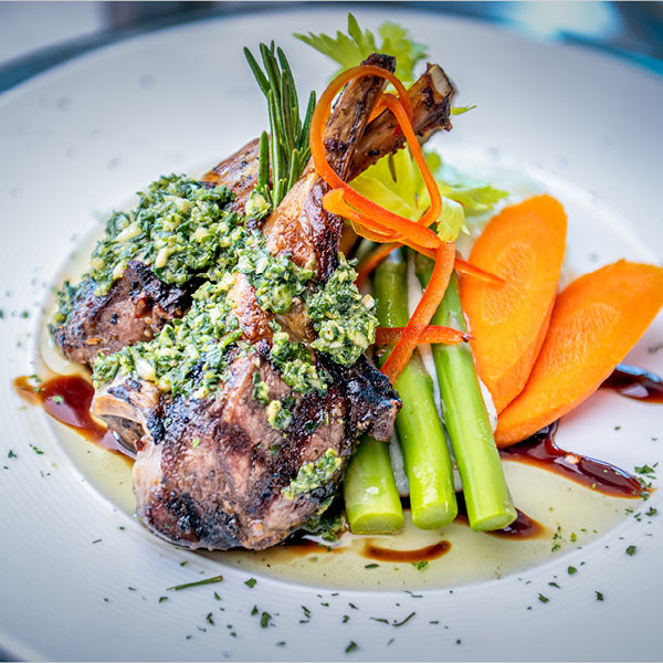 A gourmet dish featuring grilled lamb chops topped with herb pesto is served with steamed asparagus, carrot slices, and garnished with rosemary, thin carrot strips, and green herbs on a white plate. A streak of brown sauce decorates the plate next to the vegetables.