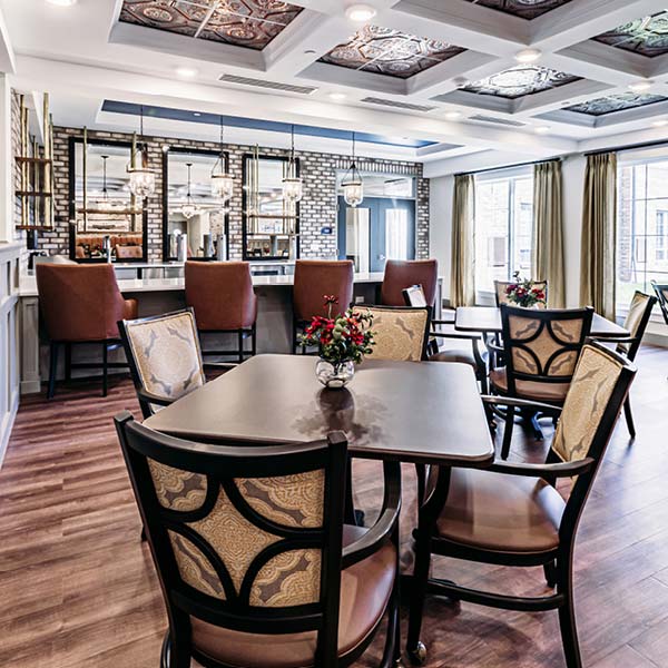 A neatly arranged dining area featuring a brown wooden floor, square tables with upholstered chairs, and decorative ceiling panels. The space is accentuated by hanging lights, a brick wall, large windows with curtains, and a bar area with cushioned high chairs.