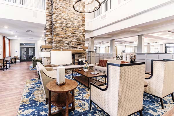 A spacious, modern lounge area with high ceilings, featuring a central stone fireplace, comfortable seating including sofas and armchairs, and a large blue-patterned rug. The room has a mix of natural and artificial lighting, with tables and decor accents.
