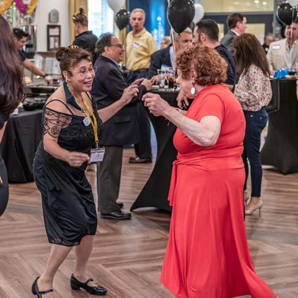 Two women are dancing together at a lively indoor event. The woman on the left is dressed in a black dress with lace details, and the woman on the right is wearing a bright orange dress. People in the background are mingling and holding balloons. The atmosphere is festive.