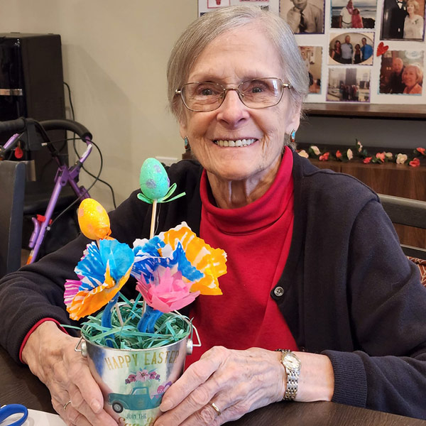 An elderly woman with glasses and short gray hair smiles while holding a colorful Easter-themed craft. The craft features eggs and flowers in a small bucket labeled "Happy Easter." She is wearing a red turtleneck with a dark cardigan, sitting at a table.