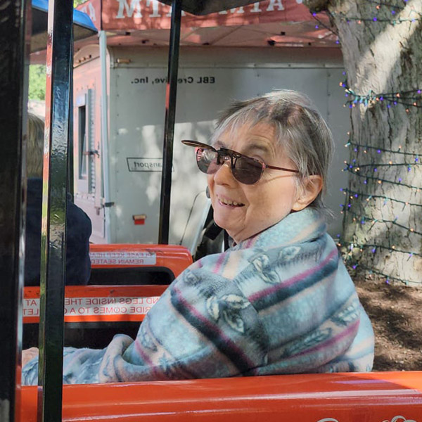 A woman with short gray hair is smiling while sitting in a small outdoor train ride. She is wearing sunglasses and a light blue patterned blanket over her shoulders. In the background, there is a trailer and a tree wrapped with string lights.