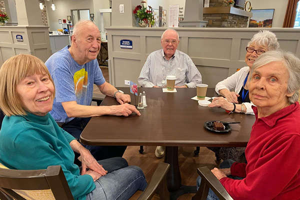 Five elderly individuals are sitting around a table in a communal space. Three men and two women are smiling at the camera. They have drinks and snacks in front of them and appear to be enjoying each other's company in a bright, welcoming environment.