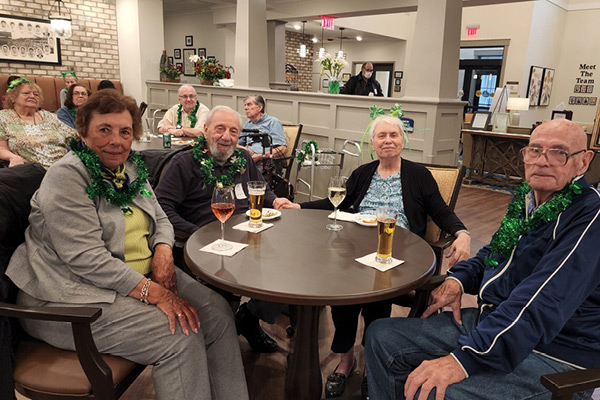 A group of elderly people with green tinsel leis around their necks sit around a table in a communal area, enjoying drinks. They appear to be celebrating an event. The background shows more people sitting and talking in a warmly lit room.