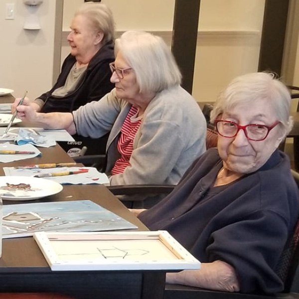 Three elderly women are sitting at a table engaged in painting activities. The woman on the right, wearing red glasses, looks directly at the camera with a slight smile. The women are surrounded by art supplies, including plates with paint and brushes.