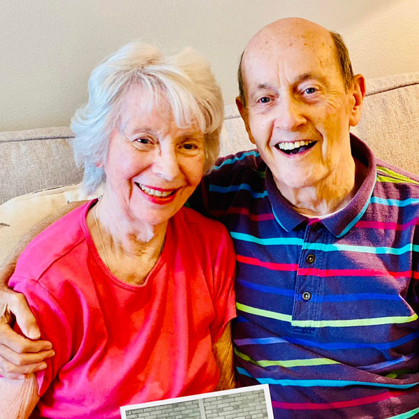 An elderly couple, both smiling brightly, sits close together on a couch. The woman, with short white hair, is wearing a pink shirt, and the man, with thinning hair, is wearing a colorful striped polo shirt. They appear happy and affectionate, with the man's arm around the woman.