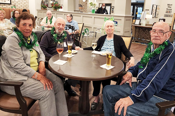 Four elderly people, two women and two men, are sitting around a round table in a communal area, each wearing green garlands. They have drinks in front of them, and other elderly individuals are visible in the background. The setting appears to be festive and social.
