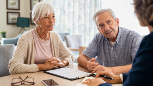 An elderly couple is seated at a table, listening attentively to a professional who is gesturing with a pen. The man and woman have a document in front of them, alongside a cellphone. The room is well-lit, with a cozy and professional atmosphere.